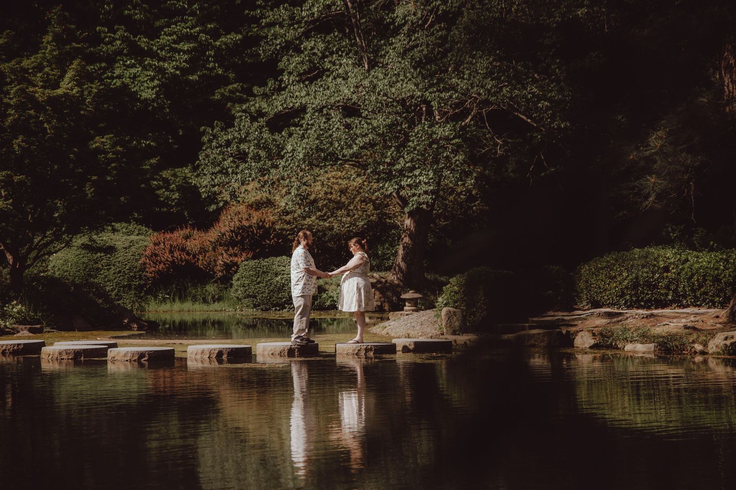 Couple holding hands on stepping stones over a tranquil pond in a lush green garden setting.