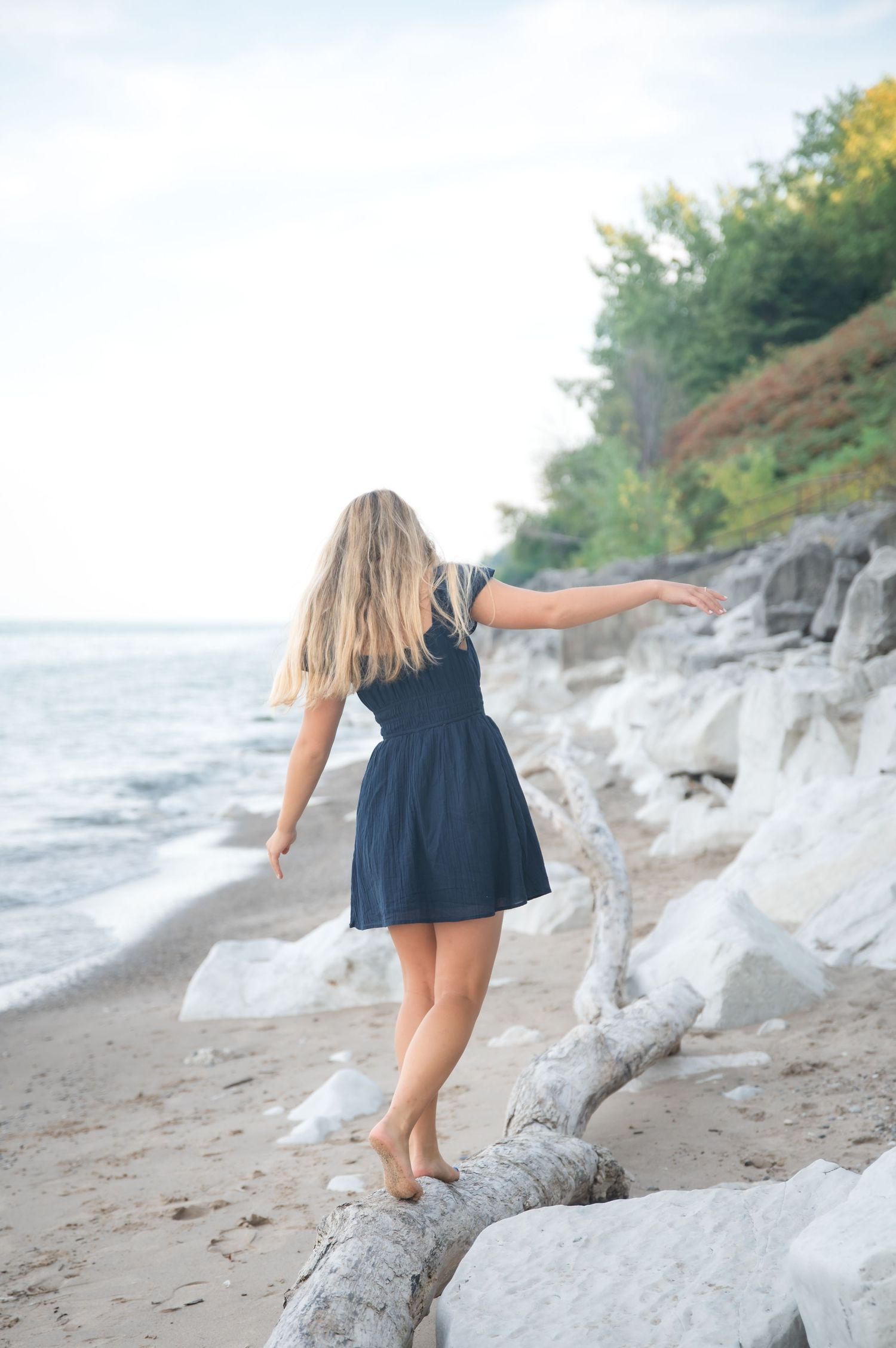 Blonde woman in navy dress balancing on driftwood log on rocky beach shoreline with trees in background.