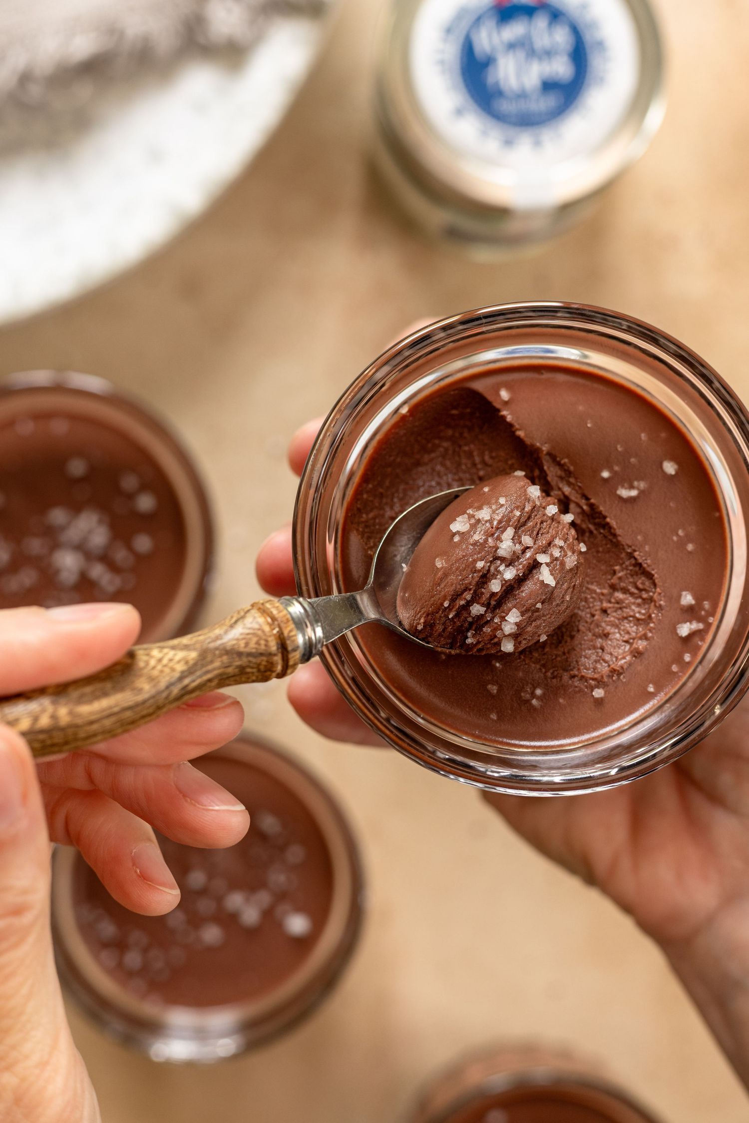 Hands holding a glass jar of rich chocolate mousse topped with flaky sea salt, with a spoon scooping into it.