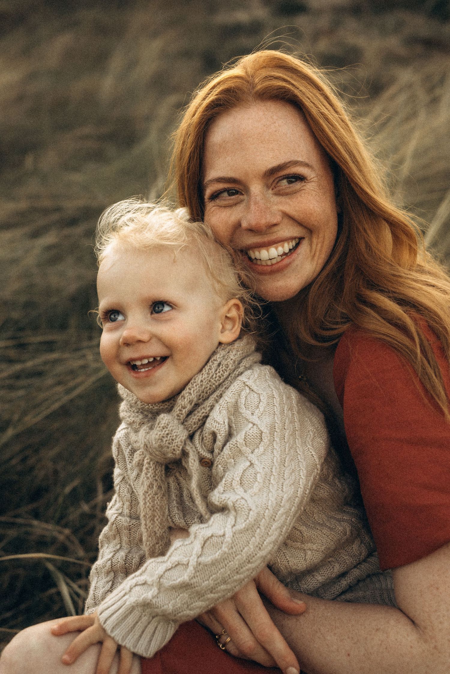 Smiling redhead mother hugging her happy blonde toddler outdoors in a grassy field, both wearing cozy knit sweaters Javea photographer