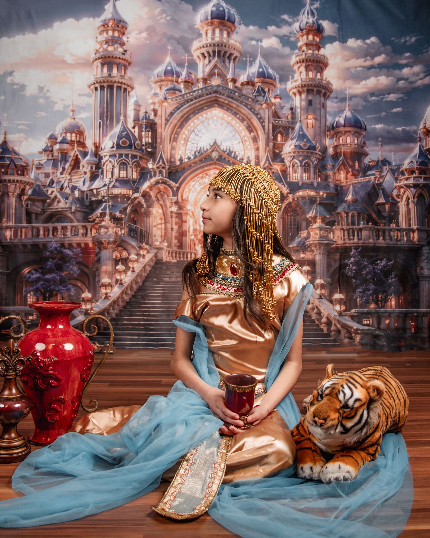 Young girl dressed as Cinderella in blue gown with pearl necklace, posing before a magical pumpkin carriage backdrop.