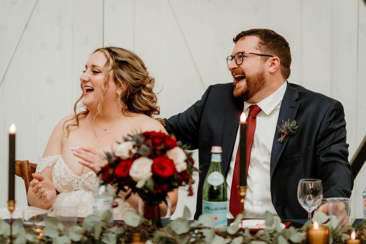 Bride and groom laughing joyfully at wedding reception head table with red rose bouquet and candles.