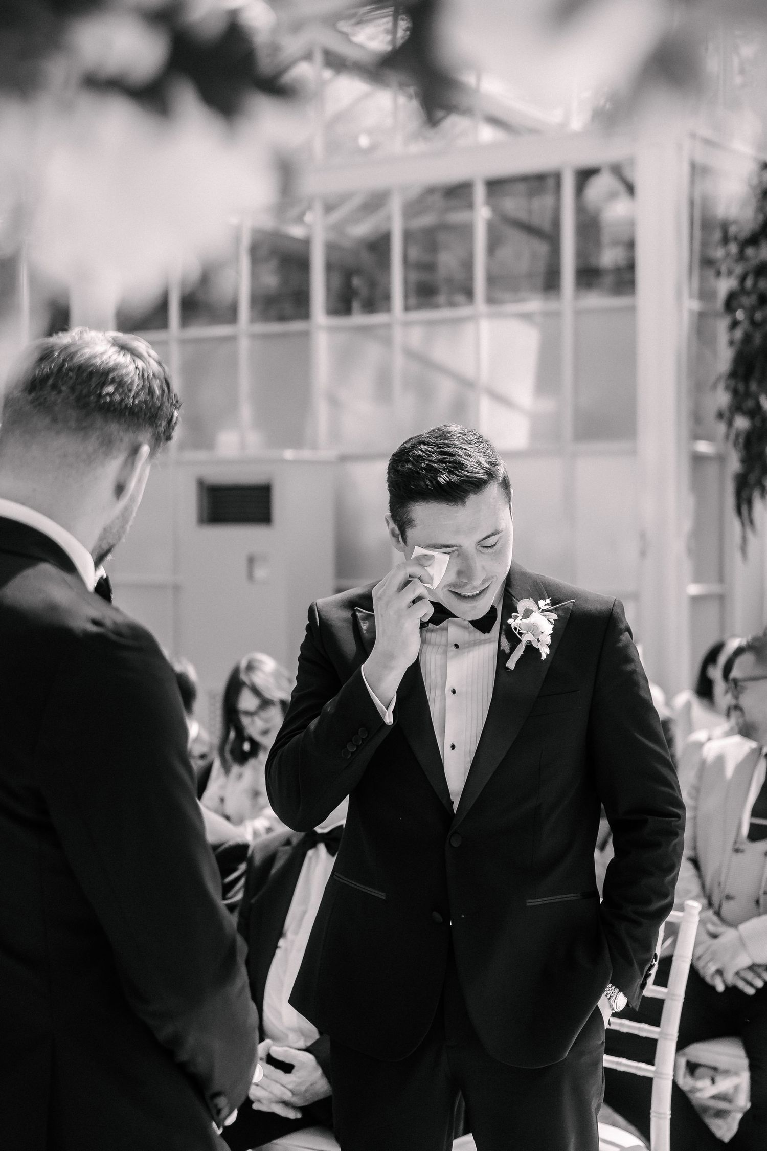 Emotional groom wiping tears at wedding ceremony in black and white photo, wearing tuxedo with boutonniere.