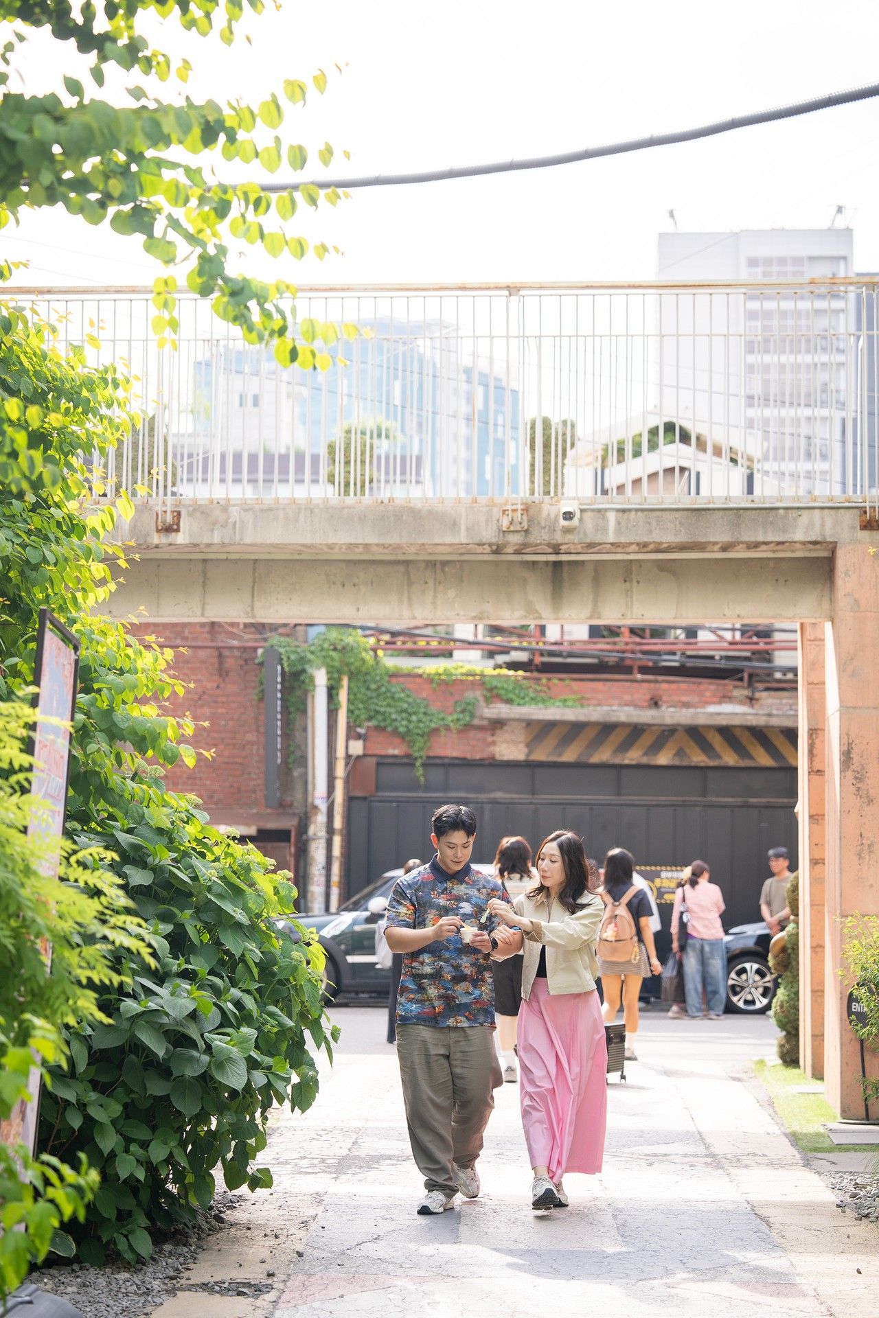A couple walks together along a tree-lined urban pathway beneath a concrete bridge in a city setting.