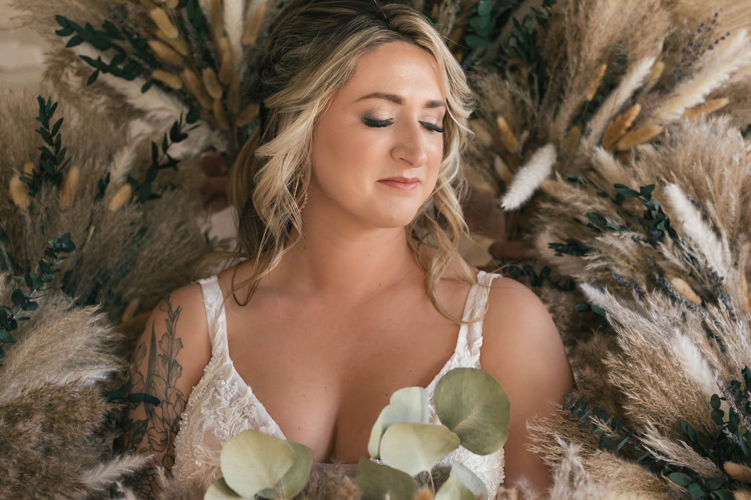 Bride lying among dried pampas grass and greenery holding eucalyptus in a boho bridal portrait.
