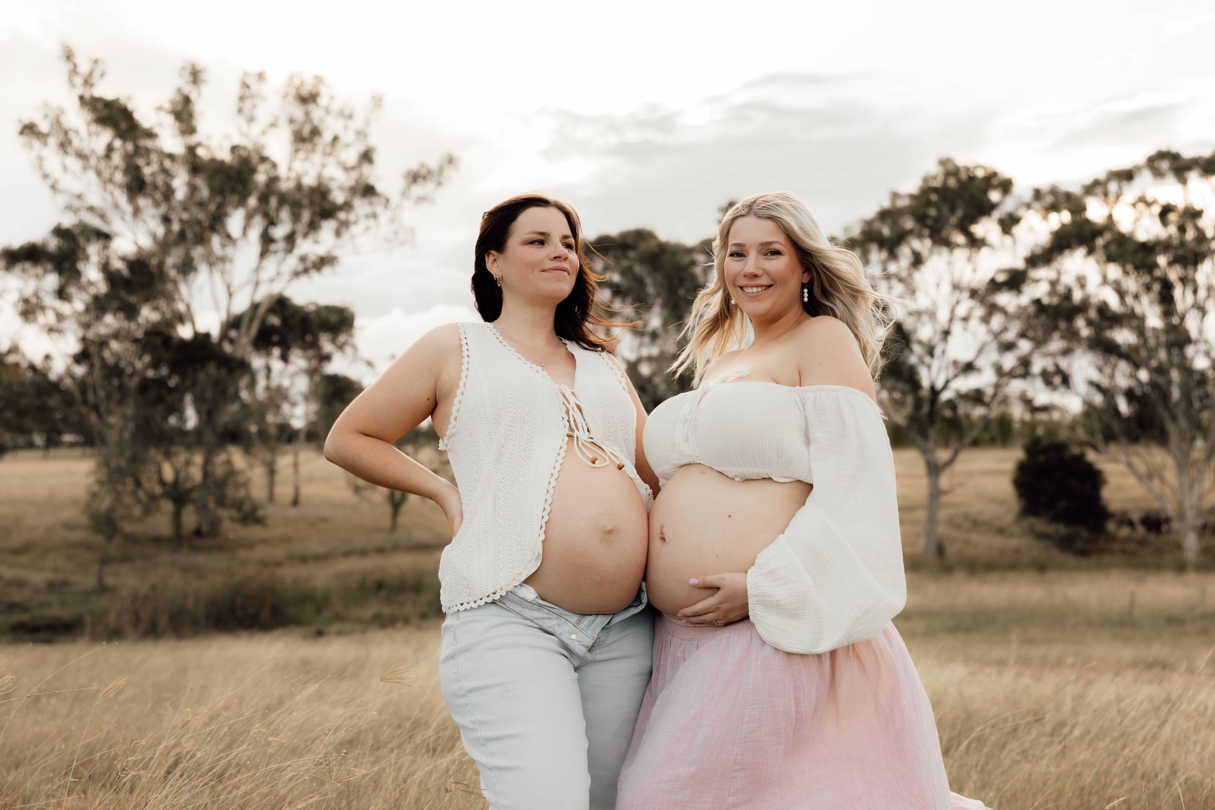 Two pregnant women pose together in a golden field at sunset, wearing white tops and pastel skirts.