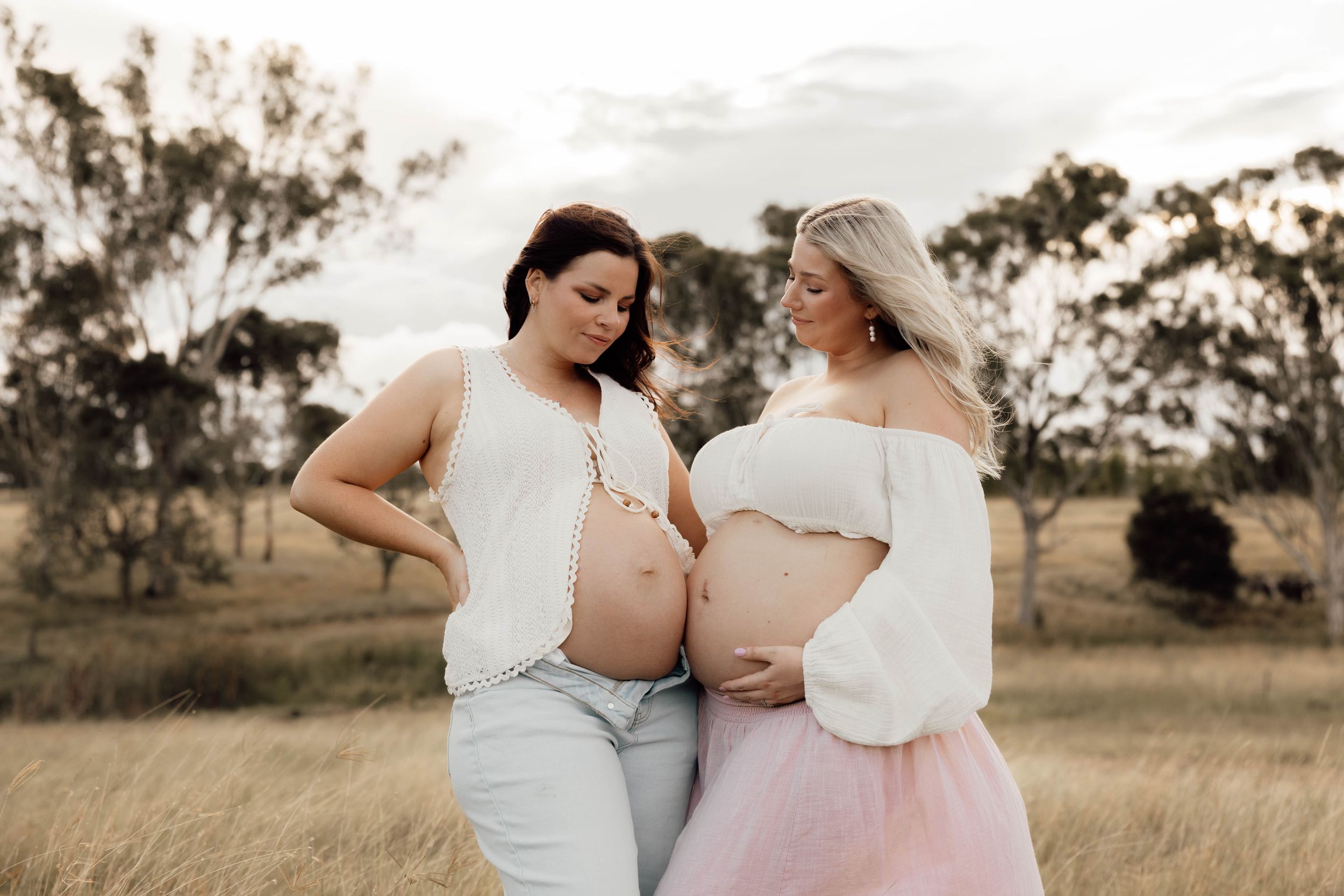 Two pregnant women in white tops pose together in a golden field at sunset during a maternity photoshoot.