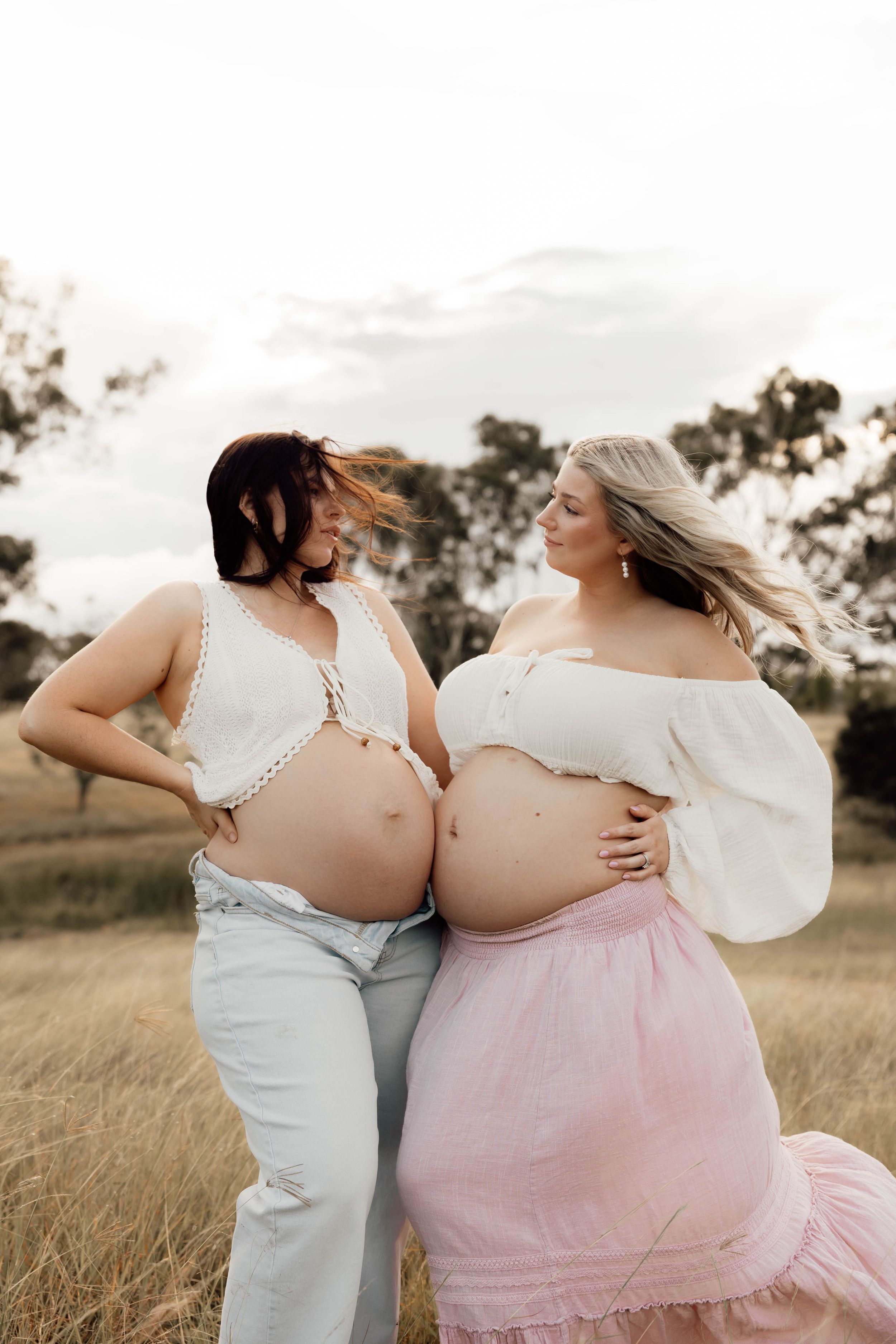 Two pregnant women in white tops pose together in a golden field, showcasing their baby bumps in a maternity photoshoot.