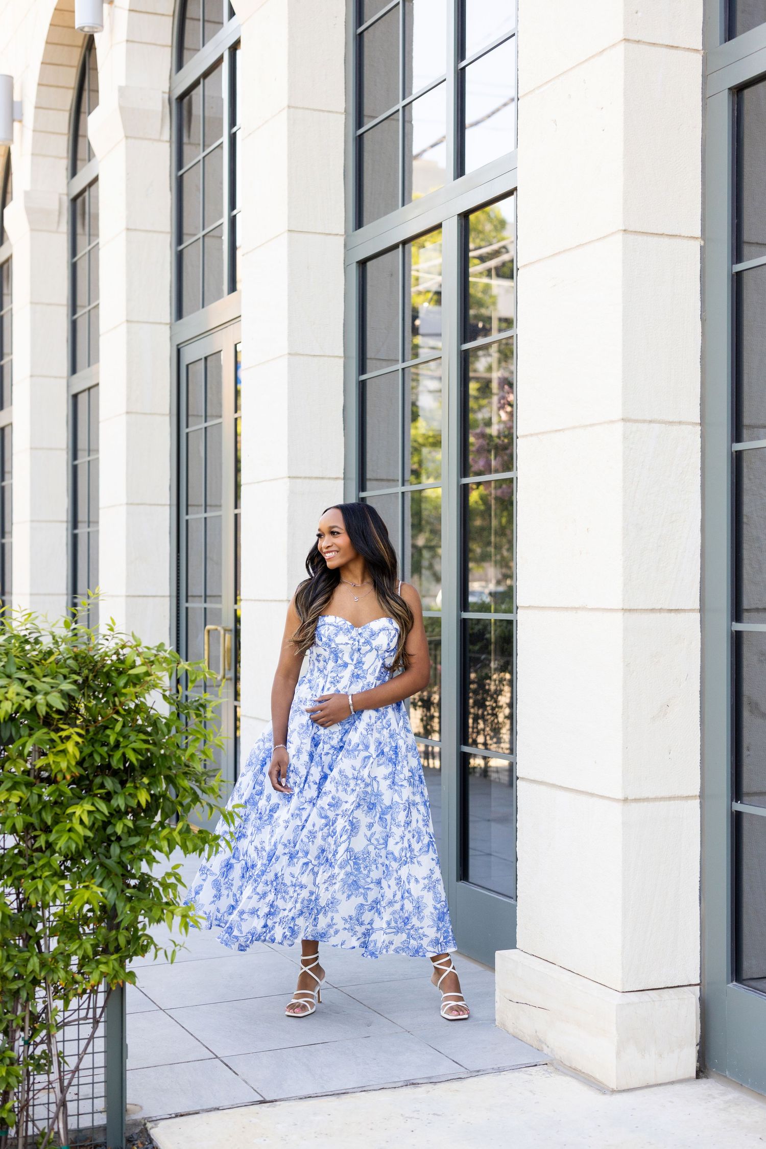 Woman in blue floral maxi dress and strappy heels posing outdoors near elegant building entrance with greenery.