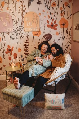 A couple sits in a cozy nursery chair, lovingly gazing at their newborn baby, surrounded by floral wallpaper.