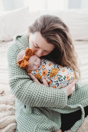 Mother tenderly kissing sleeping newborn wrapped in floral swaddle with orange bow headband.