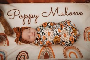 Newborn baby named Poppy Malone swaddled in floral wrap with rust bow on rainbow blanket.