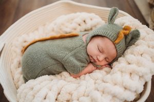 Sleeping newborn baby in green knit bunny hat and outfit nestled in white woven basket.