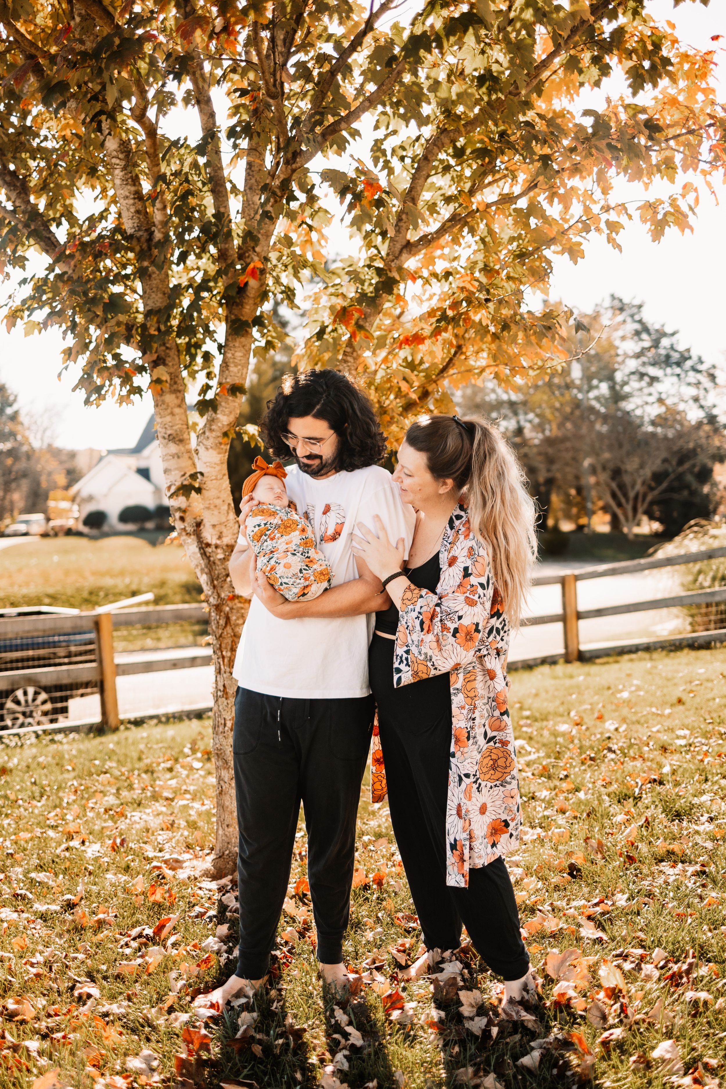 Young couple holding newborn baby under autumn tree with colorful fall foliage on a sunny day outdoors.