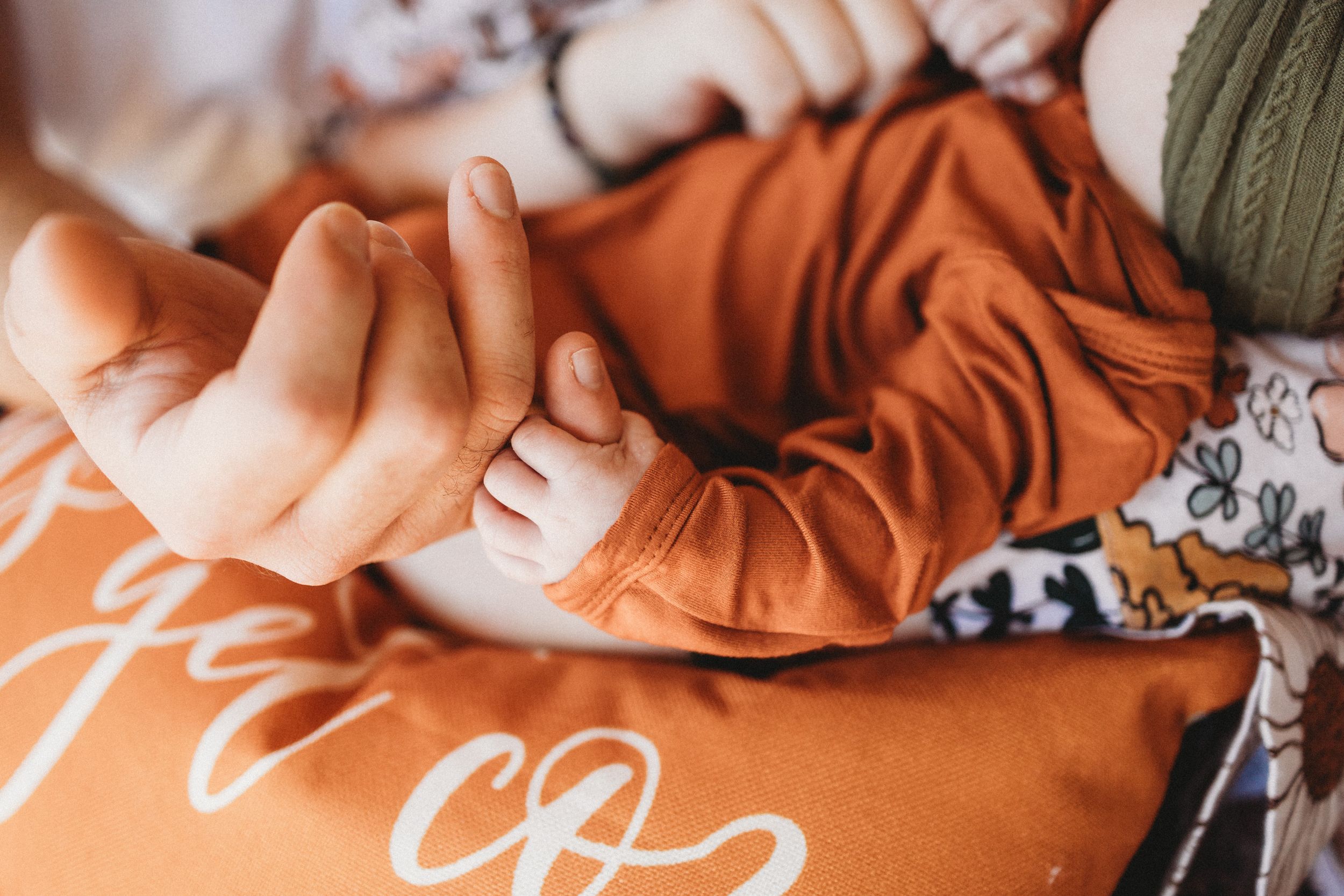 Newborn baby grasping adult finger while wearing rust orange outfit on matching pillow.