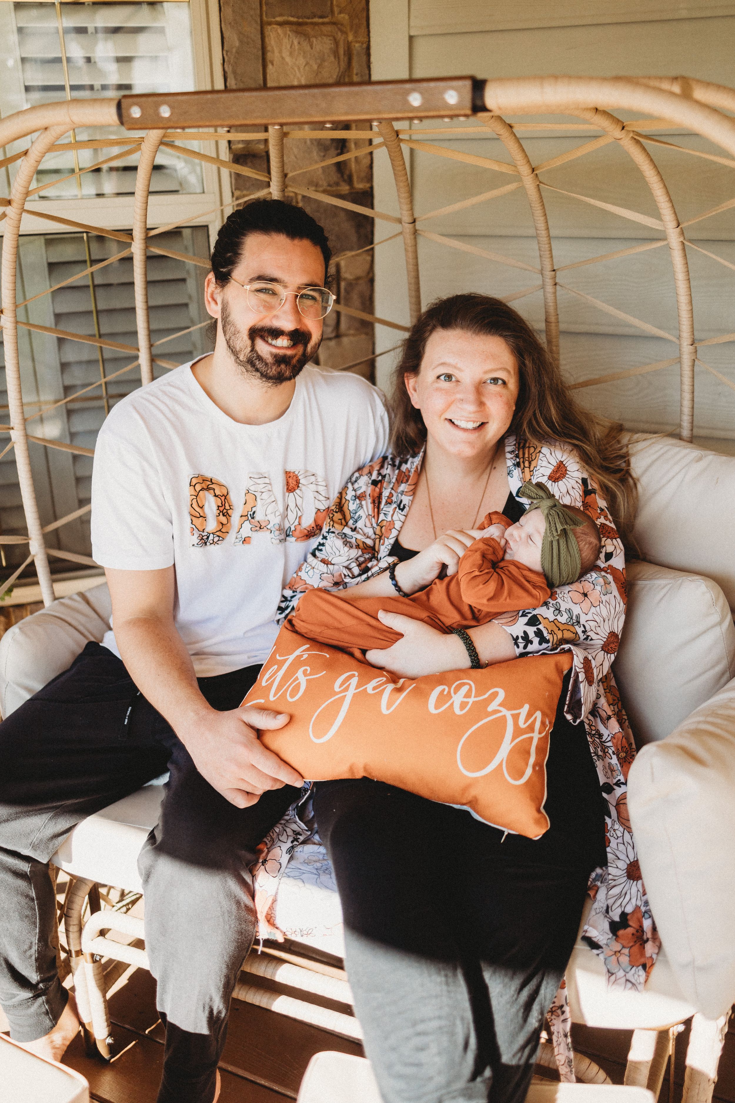 Smiling couple sitting in wicker egg chair on porch holding newborn baby and orange ‘let’s get cozy’ pillow.