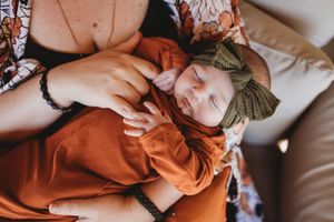 Sleeping newborn baby in orange outfit and green bow headband cradled in mother's arms.