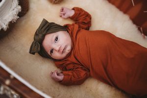 Newborn baby in rust orange outfit and olive bow headband lying in white bassinet.