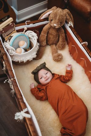 Smiling baby in rust onesie lying in wooden bassinet with fluffy sheepskin, bunny plush toy, and wicker basket.