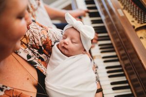Sleeping newborn baby wrapped in white swaddle with bow headband near piano keys.