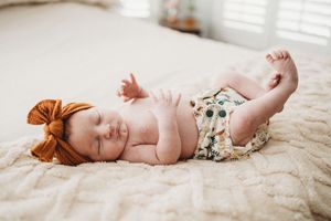 Sleeping newborn baby in floral bloomers and rust bow headband resting on cream bedding.