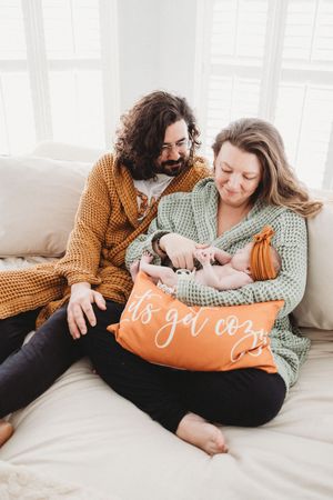 Happy couple in cozy sweaters sitting on bed admiring their newborn baby with an orange decorative pillow.