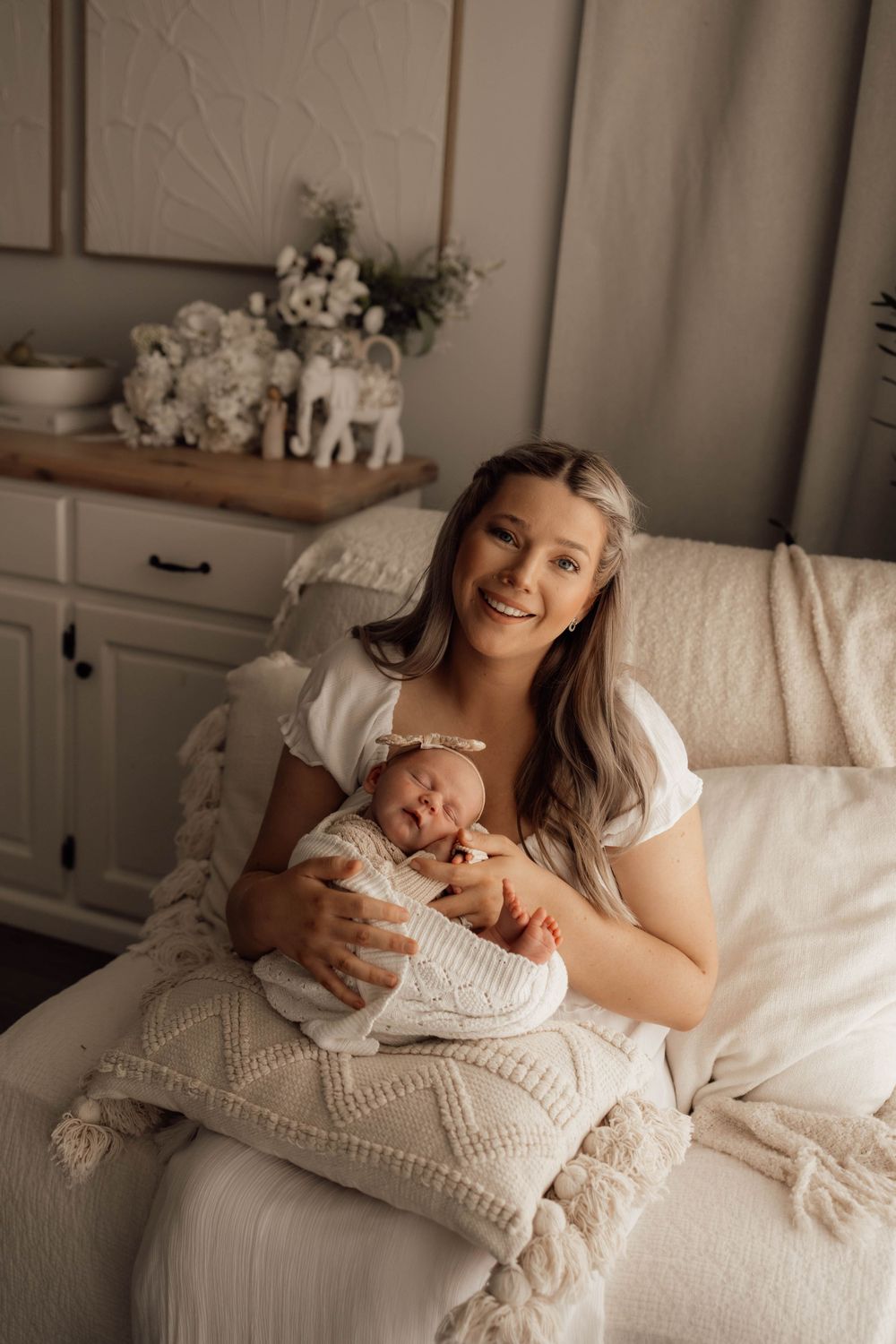 Smiling mother in white dress holding sleeping newborn baby wrapped in knit blanket on a couch.