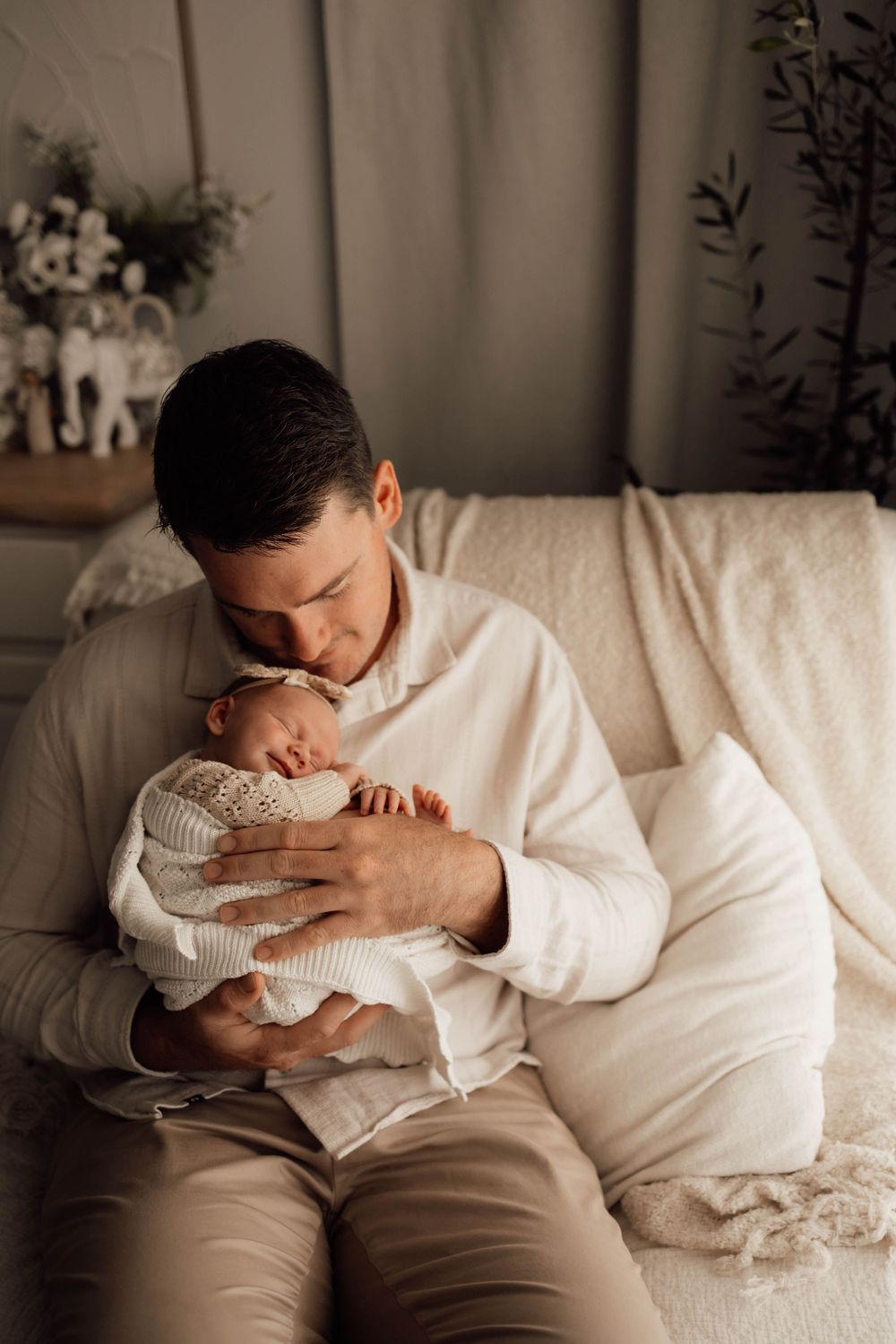 Father sitting on couch tenderly holding sleeping newborn baby dressed in white knit outfit.