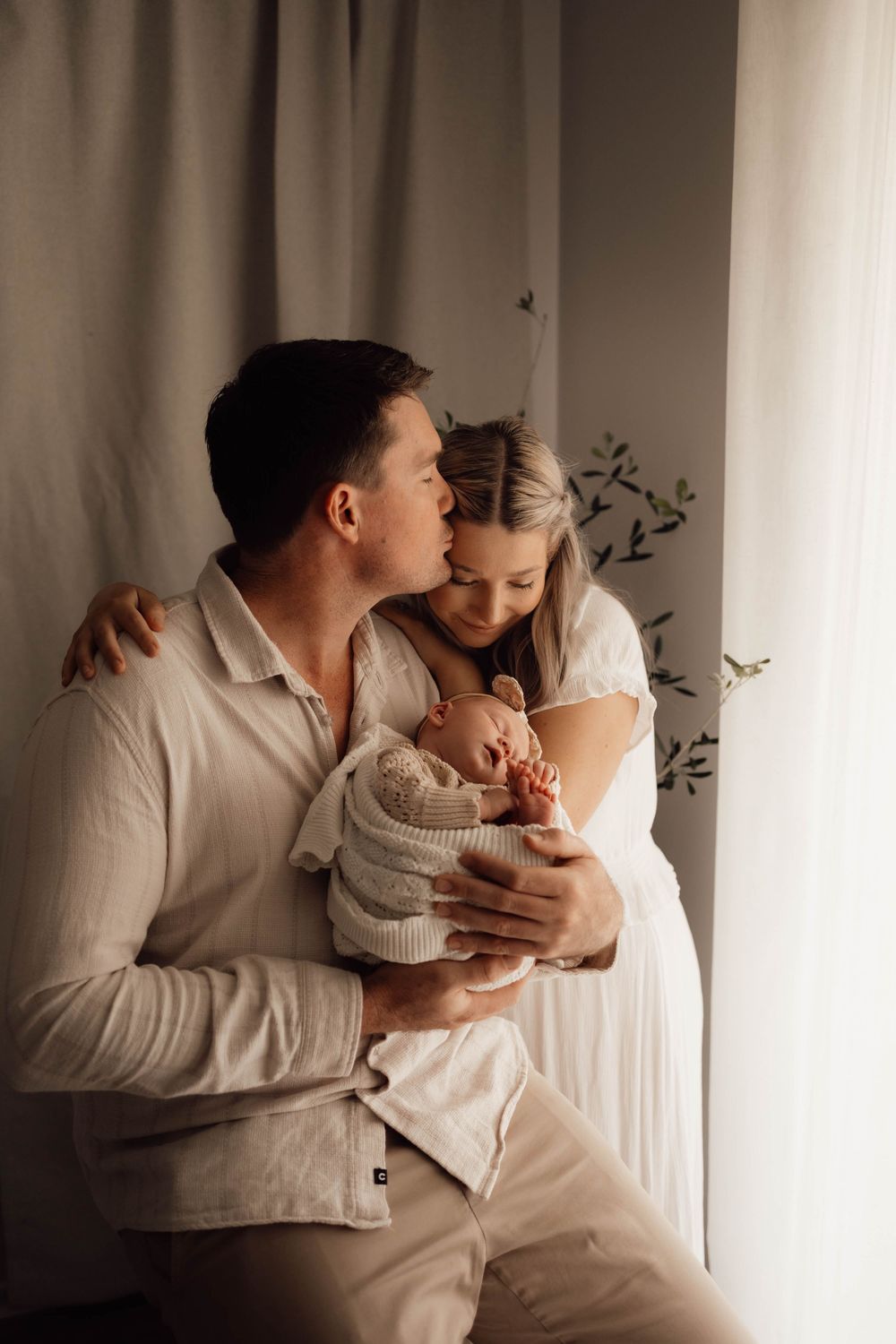 Parents in white outfits tenderly holding newborn baby by a sunlit window.