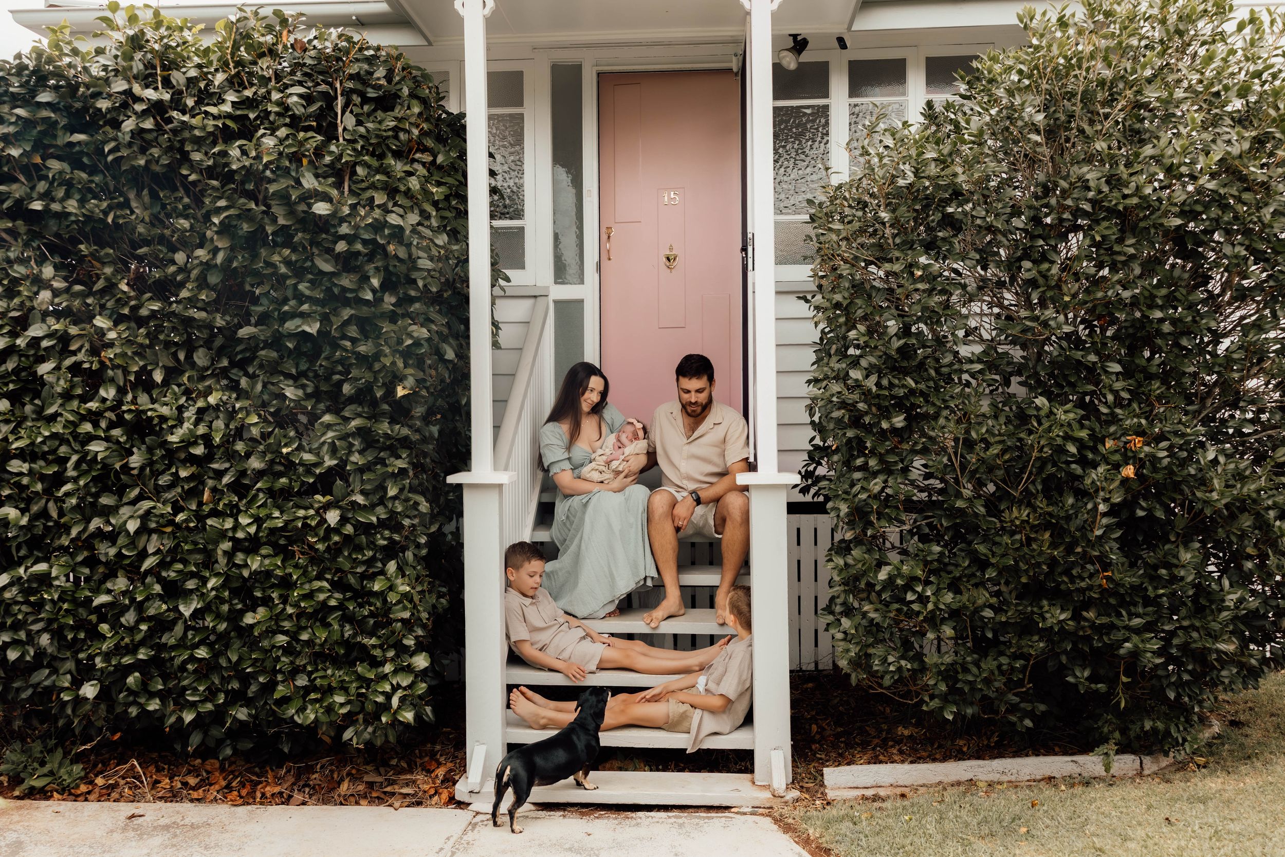 Family with newborn baby and young boy sitting on front porch of white home with dog.