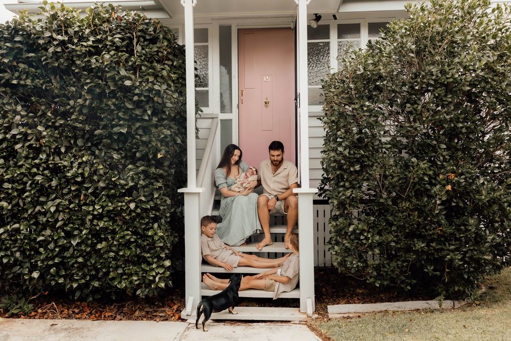 Family with newborn baby and young boy sitting on front porch of white home with dog.