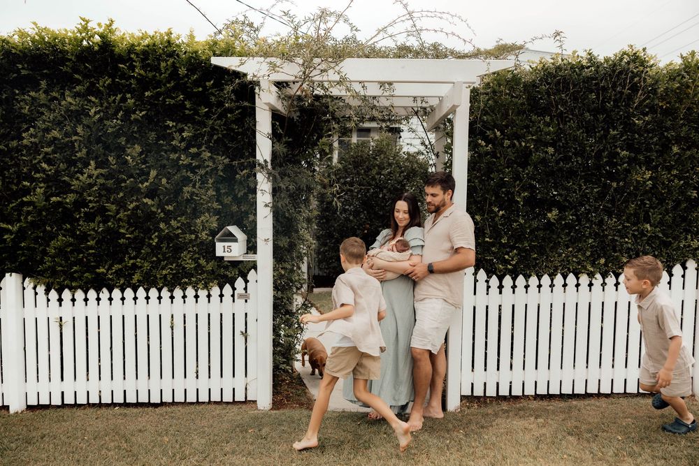 Family of four with newborn baby gathered at white picket fence garden gate, children playing.