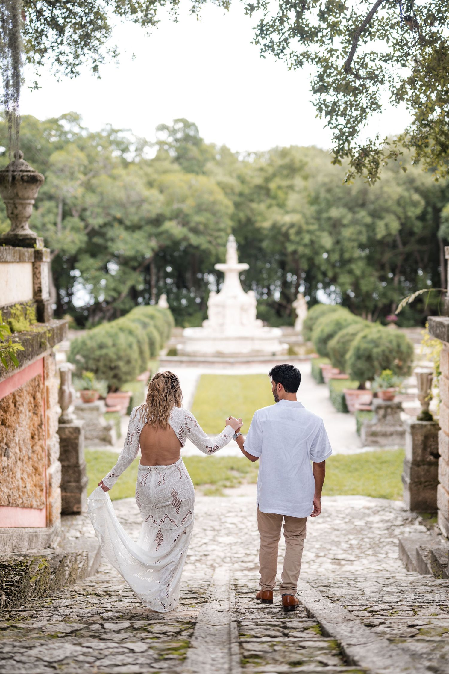 Bride and groom share a romantic dip kiss in front of a historic fountain at Vizcaya Gardens, surrounded by manicured greenery.