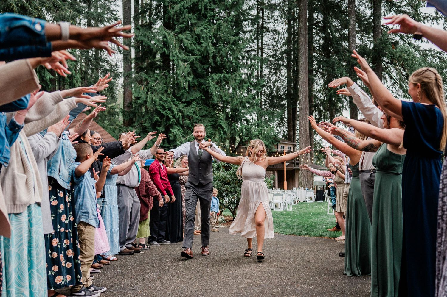 Joyful newlyweds walk through a celebratory tunnel of raised arms from guests at an outdoor forest wedding.