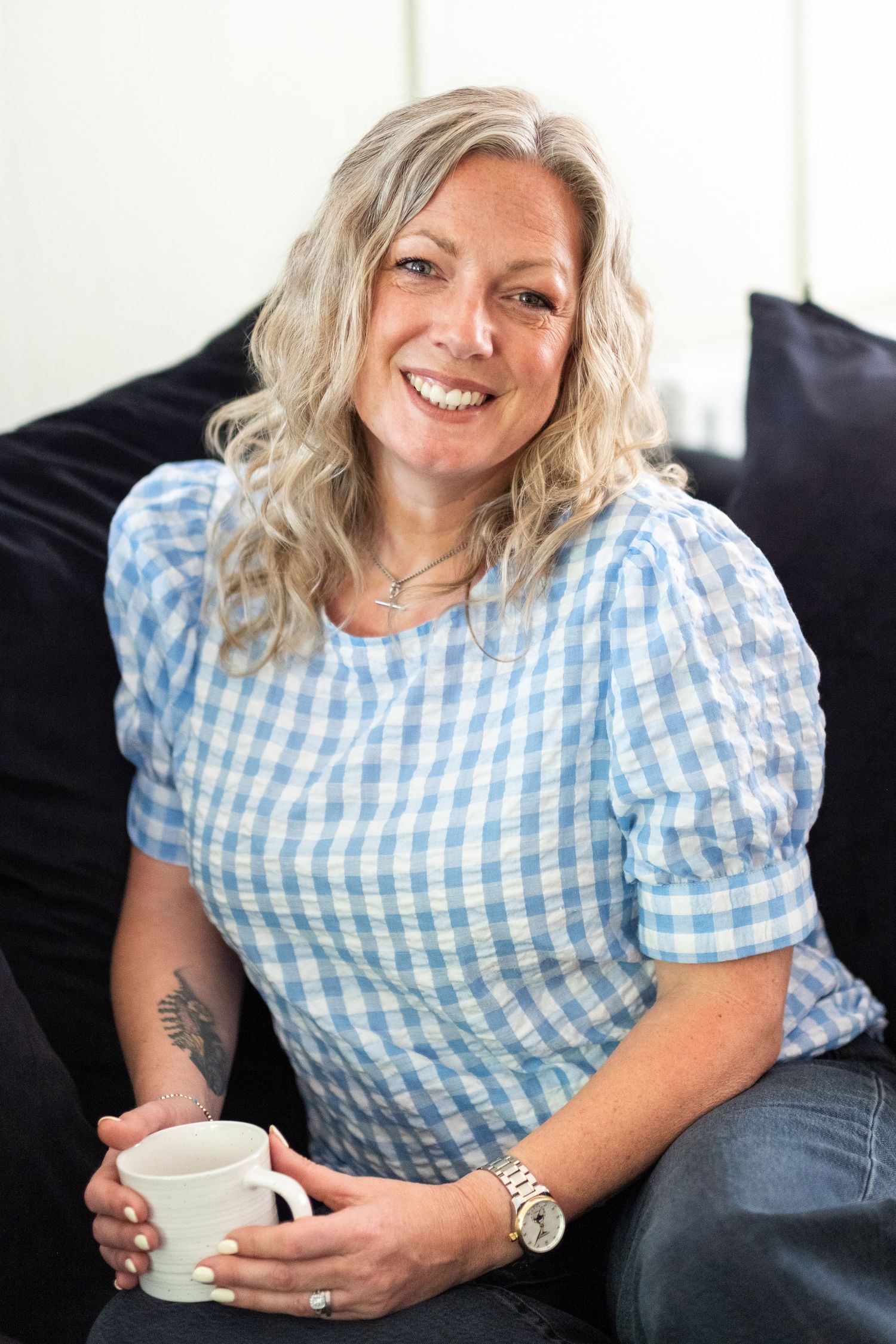 Smiling middle-aged blonde woman in blue gingham top holding a coffee mug, sitting on a dark sofa at home.