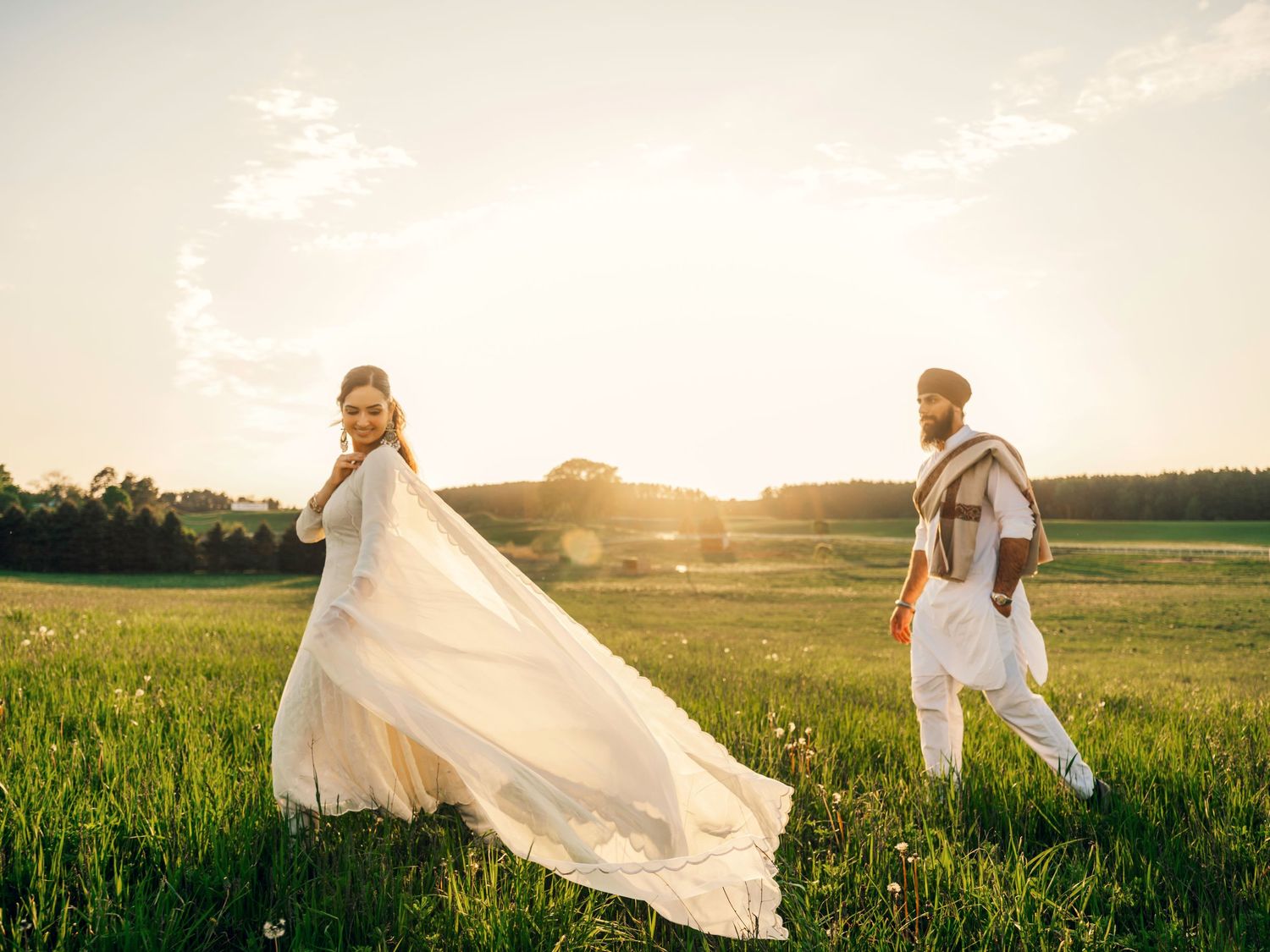 Bride in flowing white gown and groom in traditional attire stand in sunlit green field at golden hour.