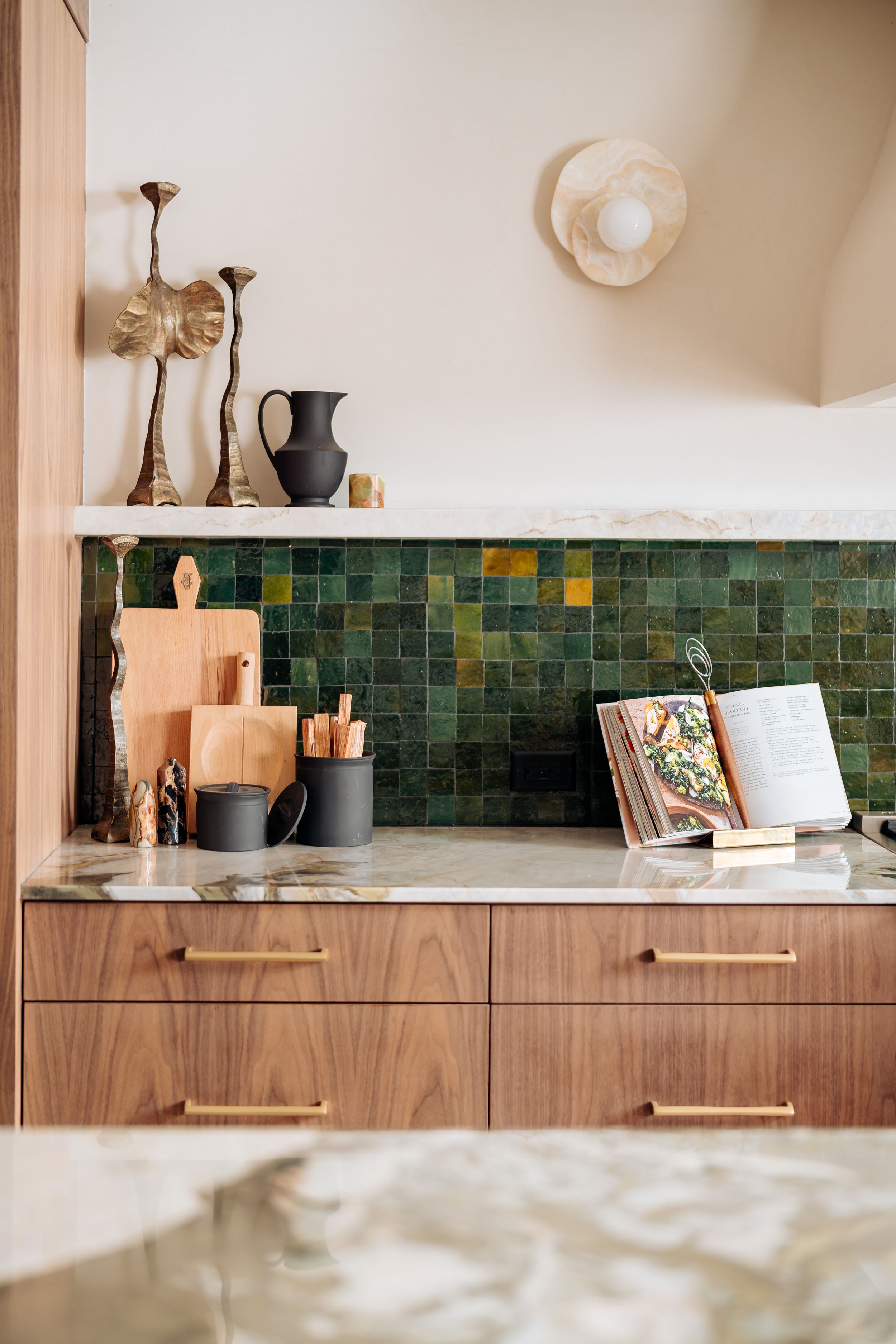 Modern kitchen with walnut cabinets, green zellige tile backsplash, marble countertop, brass hardware, and open shelf with decor.