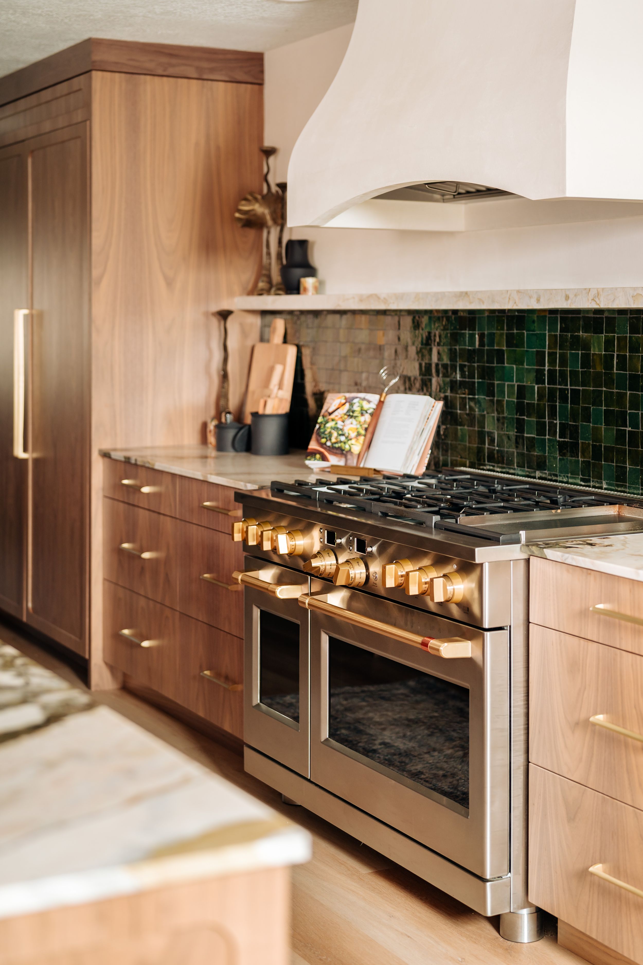 Warm walnut kitchen cabinets with gold hardware, green tile backsplash, and stainless steel dual-oven gas range.
