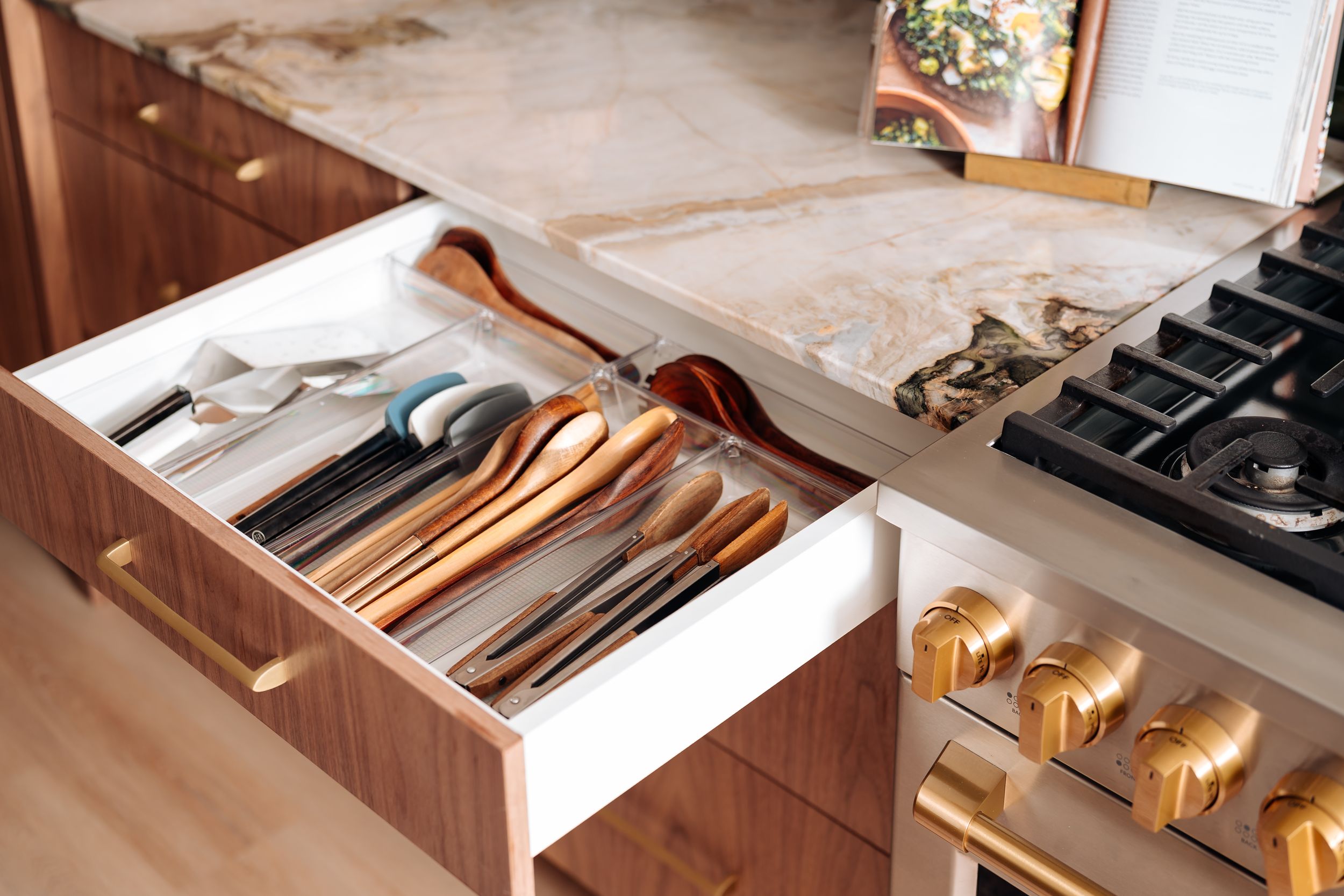 Organized kitchen drawer with wooden utensils and silverware in a divider, beside a gas stove with gold hardware.