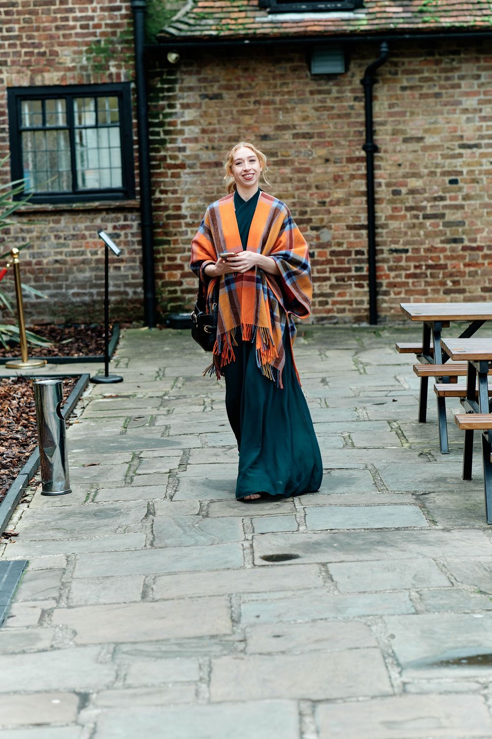 Woman in teal maxi skirt and orange plaid poncho walking on stone patio near brick building.