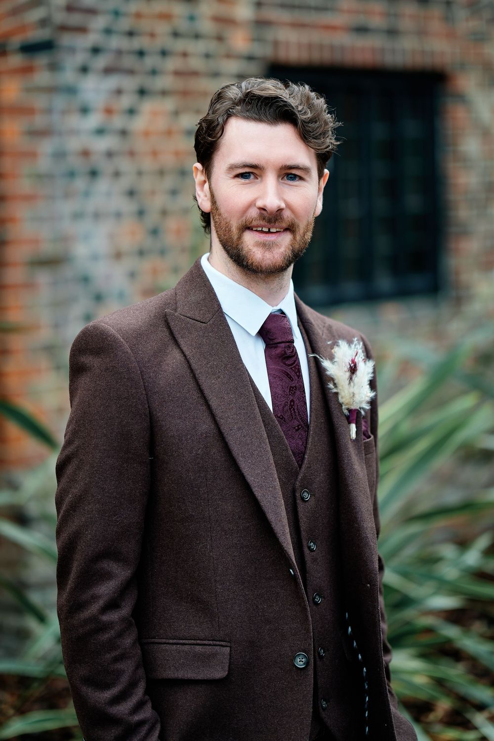 A groom in a brown tweed three-piece suit with a burgundy tie and floral boutonniere smiles outdoors near a brick wall.