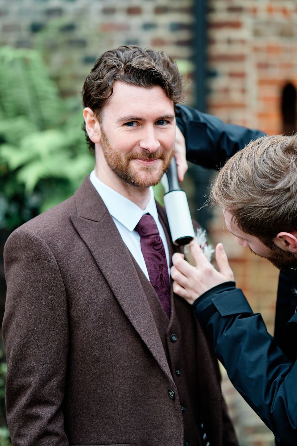 A man in a brown tweed suit and burgundy tie smiles as another person uses a lint roller on his shoulder.