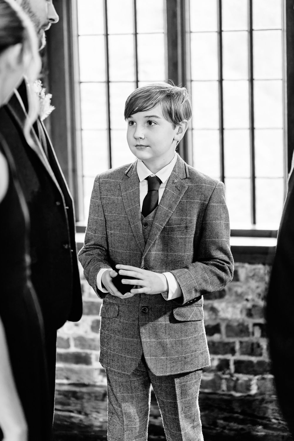 A young boy in a plaid suit and tie stands attentively at a formal event in a black and white photo.