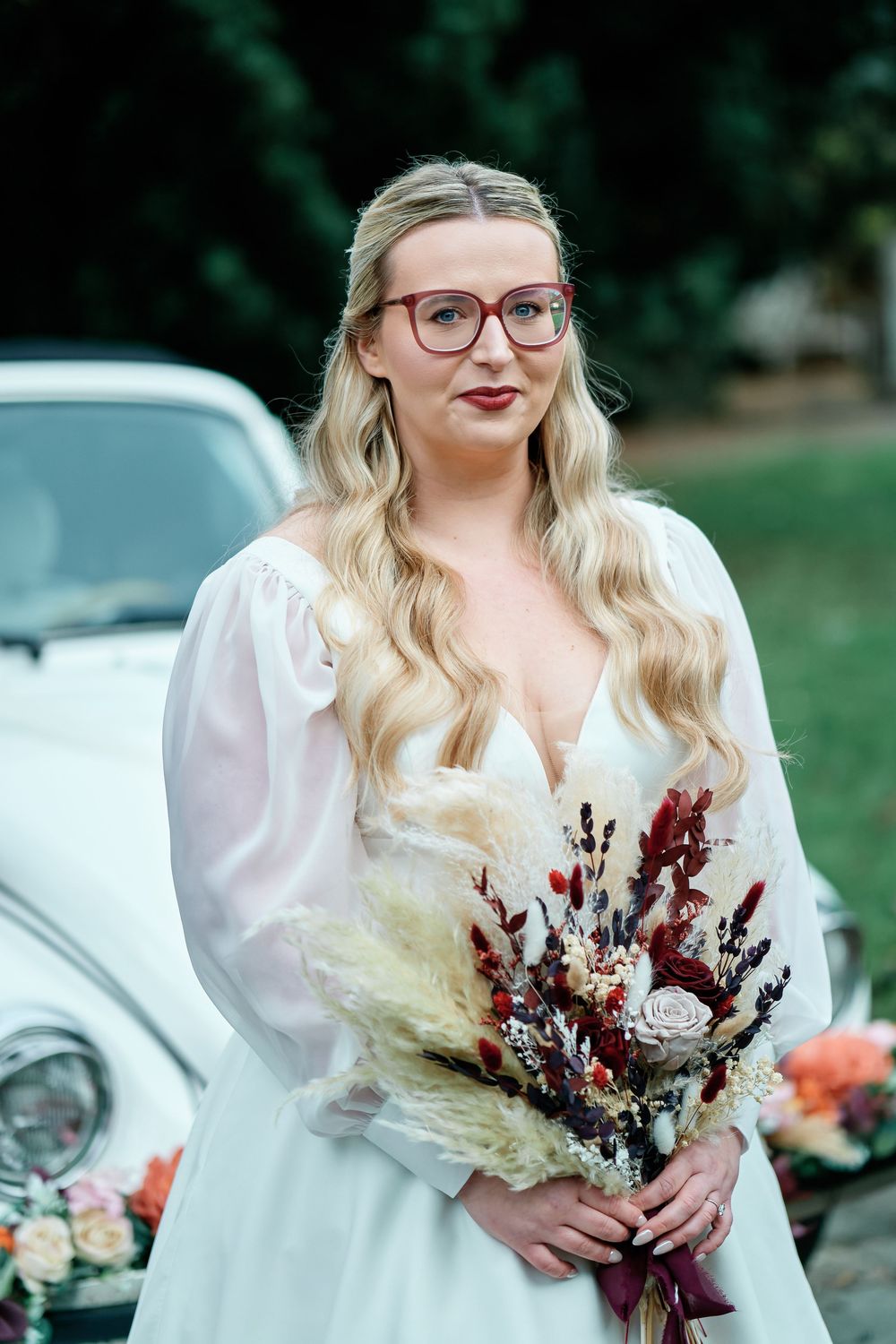 Blonde bride in white dress and red glasses holding a dried floral bouquet, standing in front of a vintage white car.