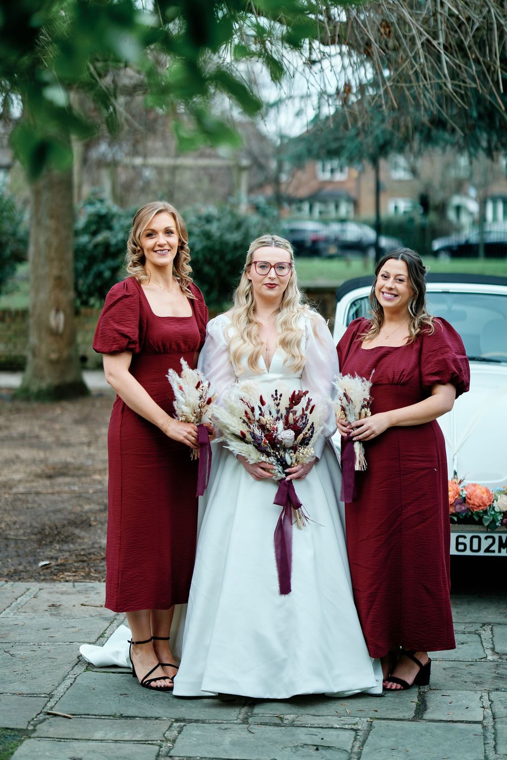 Bride in white gown with two bridesmaids in burgundy dresses holding dried floral bouquets outdoors.