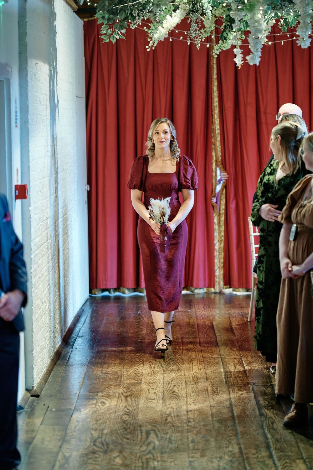 Bridesmaid walks down the aisle in a burgundy satin midi dress holding a bouquet at an elegant wedding ceremony.