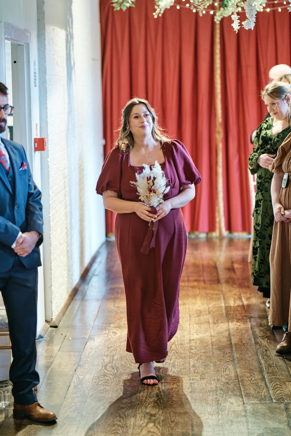 A bridesmaid in a burgundy dress walks down the aisle holding a bouquet at an elegant wedding ceremony.