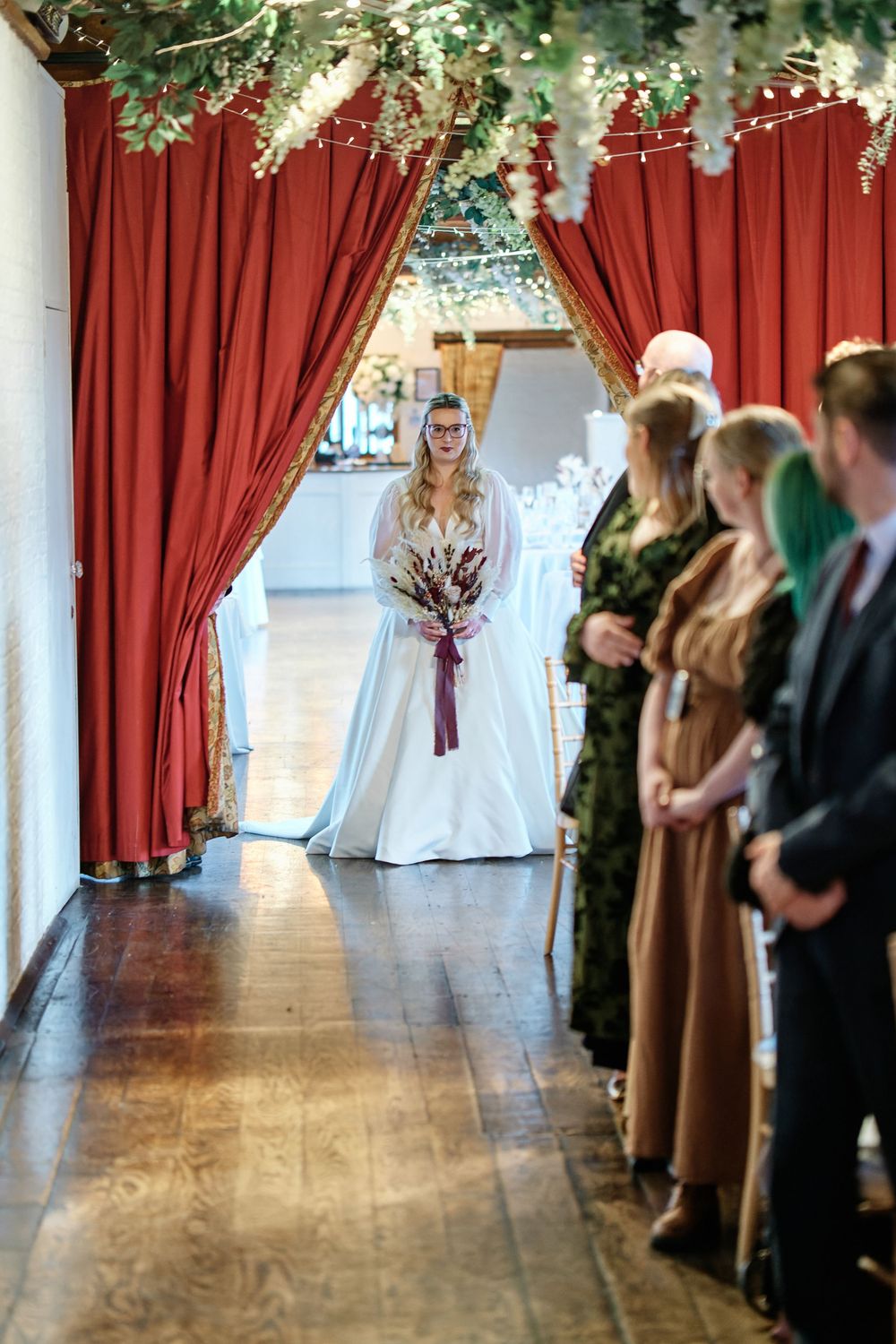 Bride in white gown holding bouquet walks down aisle between red curtains at elegant indoor wedding ceremony.