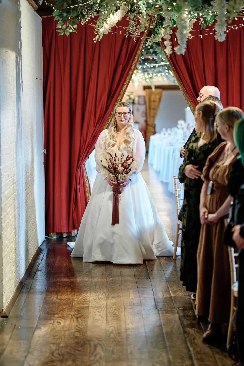 Bride in white gown holding red bouquet walks through red curtains at elegant indoor wedding ceremony.