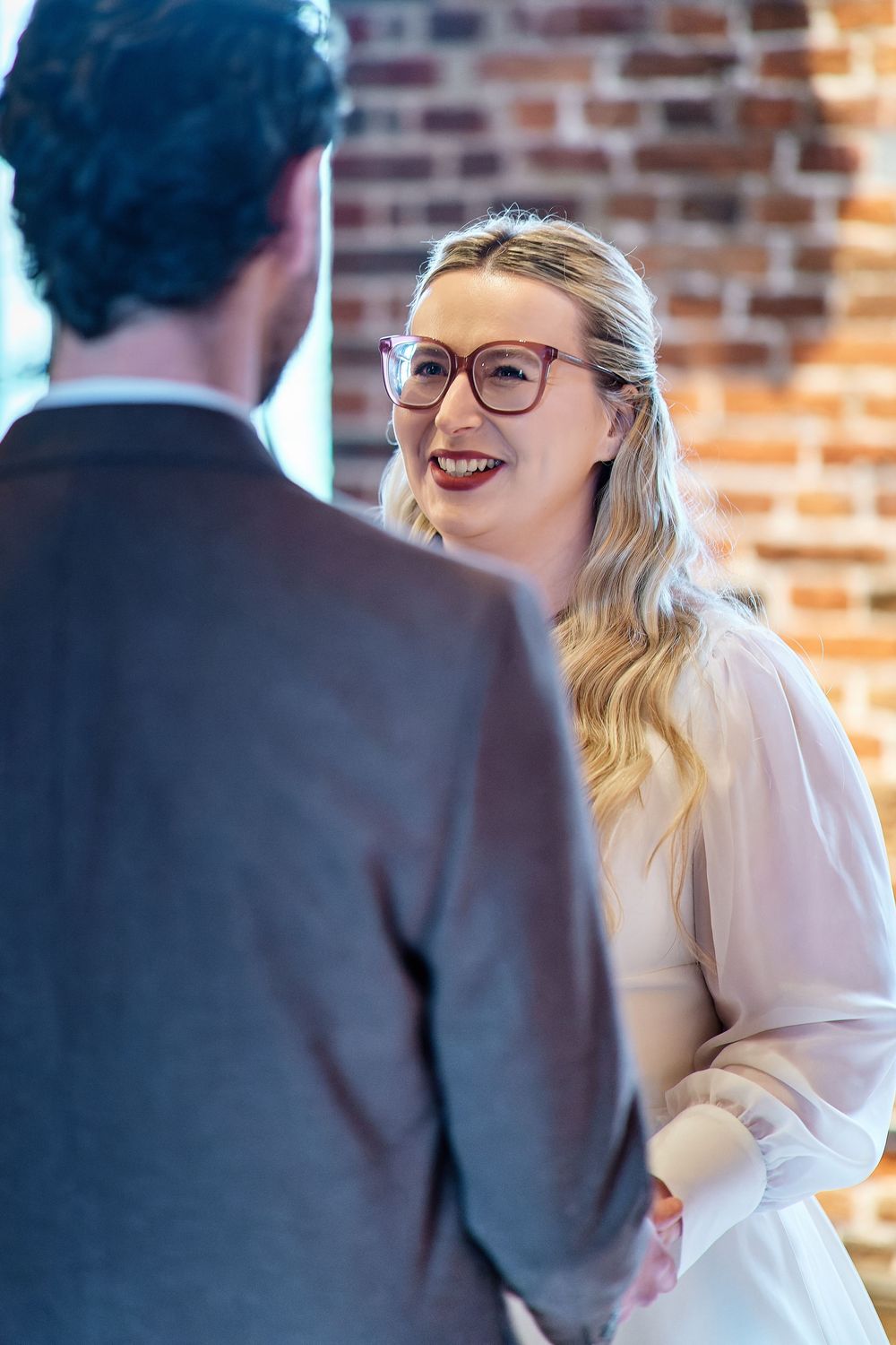Smiling blonde woman in glasses and white dress talking with a man in a suit near a brick wall.
